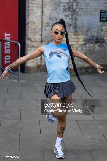 Coi Leray wears grey pleated mini skirt, blue sleeveless top with dolphine print, white socks, sneakers, sunglasses outside JW Anderson during London...