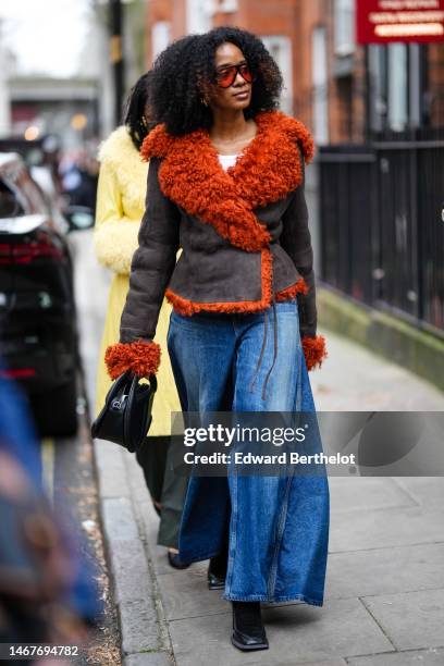 Guest wears black and red sunglasses, gold necklaces, a brown suede with orange fluffy borders jacket, a black shiny leather handbag, navy blue faded...