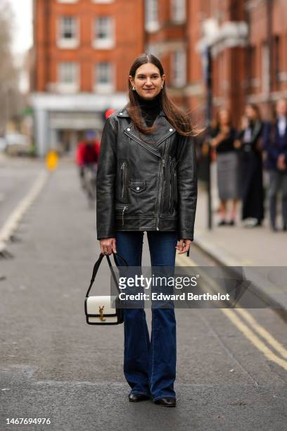 Guest wears silver earrings, a black wool turtleneck pullover, a black shiny leather zipper jacket, navy blue denim pants, a white shiny leather with...