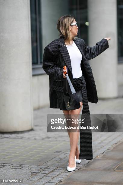 Guest wears silver sunglasses, gold and diamonds earrings, a white tank-top, a black with small white striped print pattern oversized blazer jacket,...