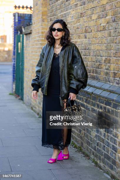 Bettina Looney wears oversized bomber jacket, black dress with slit, Prada bag, pink heeled sandals, necklace, sunglasses outside Nensi Dojaka during...