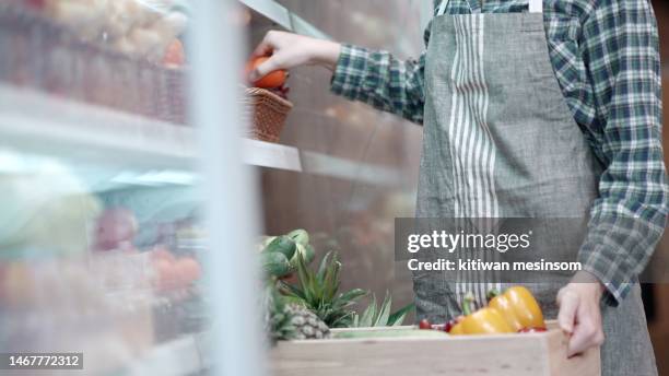 worker in apron stocking the fruits in supermarket. young employee at work, replenishes products on the shelves, works in a grocery store. female arranging vegetable and fruits on shelf an organic store. - damstrumpor bildbanksfoton och bilder