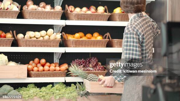 worker in apron stocking the fruits in supermarket. young employee working on fruit and vegetable department in supermarket, replenishes products on the shelves, works in a grocery store. female arranging vegetable and fruits on shelf an organic store. - stocking grocery store shelves stock pictures, royalty-free photos & images