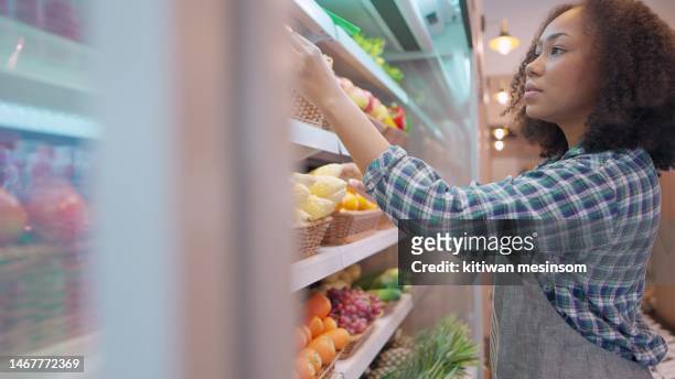 african american worker in apron stocking the fruits in supermarket. young employee at work, replenishes products on the shelves, works in a grocery store. female arranging vegetable and fruits on shelf an organic store in shopping mall. - nylon stockfoto's en -beelden