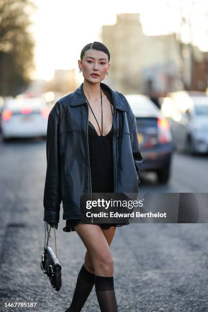 Amalie Gassmann wears silver earrings, a black halter laces neck / V-neck short dress, a black shiny leather oversized jacket, a black shiny leather...