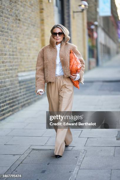 Guest wears brown sunglasses, a white shirt, a brown sheep jacket, an orange shiny leather braided large clutch, beige matte leather large pants,...