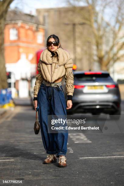 Guest wears black sunglasses, a beige quilted puffy cropped puffer jacket, a brown and beige print pattern ripped belt, navy blue denim puffy pants,...