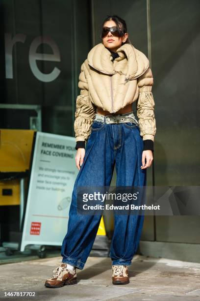 Guest wears black sunglasses, a beige quilted puffy cropped puffer jacket, a brown and beige print pattern ripped belt, navy blue denim puffy pants,...