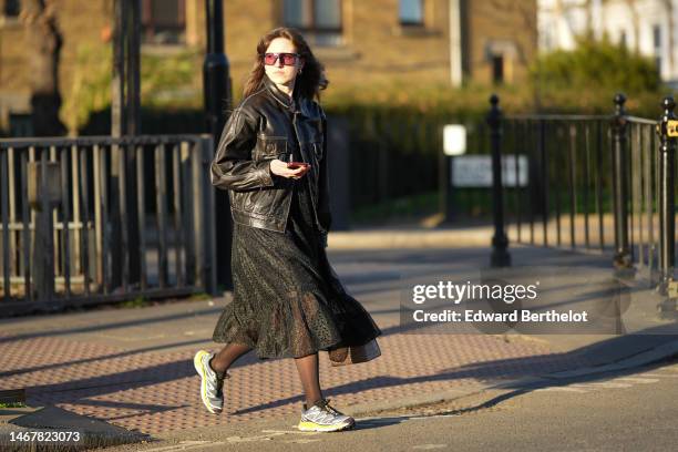 Guest wears black and red sunglasses, gold earrings, a beige with brown moon print pattern high neck t-shirt from Marine Serre, a black shiny leather...