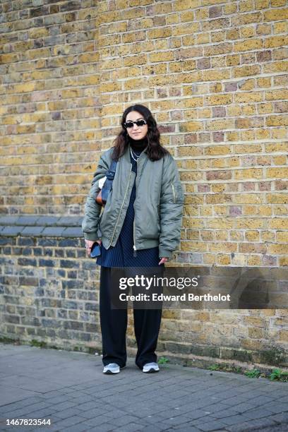 Guest wears black sunglasses, a black turtleneck pullover, gold necklaces, a khaki nylon bomber coat, a navy blue striped print pattern blazer...