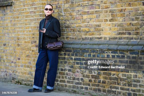 Guest wears burgundy sunglasses, gold earrings, a dark brown ribbed wool turtleneck pullover, a black blazer jacket, a dark brown shiny leather...