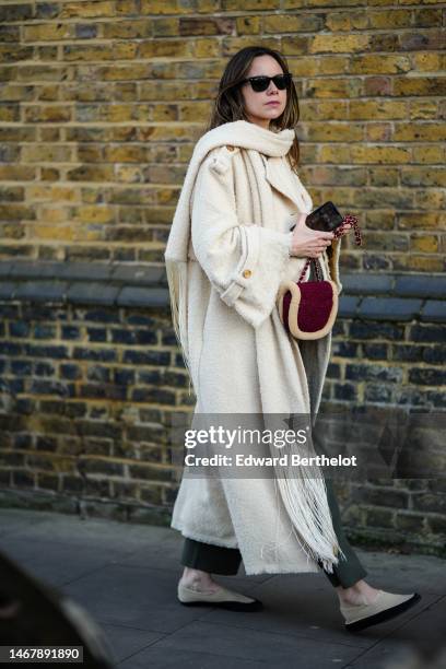 Guest wears black sunglasses, a white latte wool scarf, a white latte wool long coat, a khaki jumpsuit, a burgundy suede and white latte sheep...