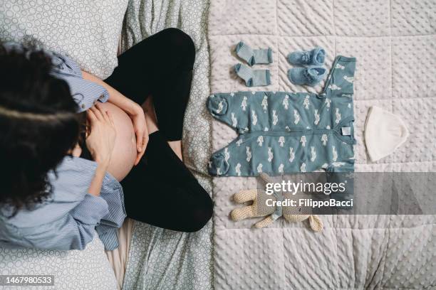 a pregnant woman is looking the clothes of her future baby on the bed - baby booties imagens e fotografias de stock