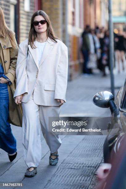 Guest wears brown sunglasses, a white t-shirt, a gold necklace, a beige oversized blazer jacket, white latte suit pants, black shiny leather nailed /...