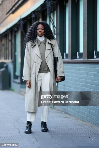 Guest wears a white wool turtleneck pullover, a khaki matte shirt, a beige long trench coat, white latte suit pants, a black shoulder bag, black...