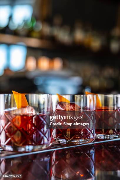 three modern glasses of negroni cocktail and gin short drink with sweet vermouth, red bitter liqueur and dried orange peel on bar counter. - cocktail counter stock pictures, royalty-free photos & images