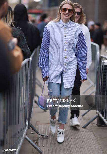 Fashion week guest seen wearing a purple and blue JW Anderson bag, oversized red shades, a striped oversized blouse, a jeans with cutouts and white...