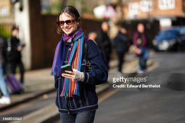 Model wears brown sunglasses, a multicolored print pattern wool long scarf, a navy blue denim buttoned jacket, dark gray denim large pants, outside...