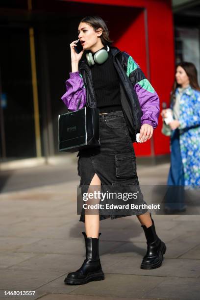 Model wears gray headphones, a black t-shirt, a black / purple / green print pattern nylon bomber coat, black denim cargo short pants, a black...