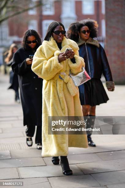 Guest wears black sunglasses, gold earrings, a black turtleneck wool pullover, a pale yellow oversized fluffy belted long coat, a silver shiny...