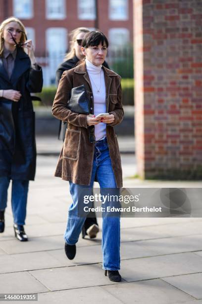 Guest wears micro earrings, a white turtleneck pullover, a brown suede long jacket, blue denim large pants, a black shiny leather clutch, black suede...