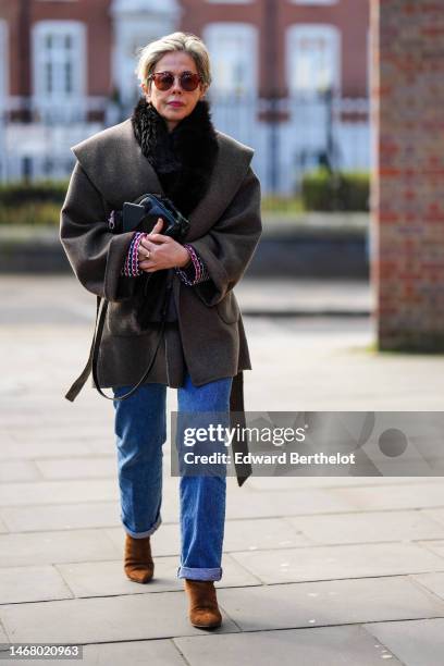 Guest wears brown sunglasses, a black fur scarf, a brown large collar belted jacket, a black shiny leather handbag, gold rings, a navy blue / red /...