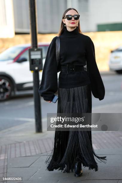 Guest wears black sunglasses, earrings, a black ribbed wool turtleneck puffy long sleeves pullover, a black long fringed skirt, black shiny leather...