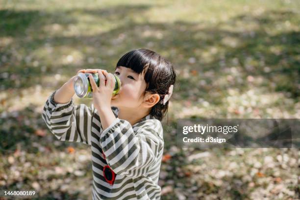 little asian girl drinking soda from can on a sunny day outdoors in the park - fizzy drink stockfoto's en -beelden