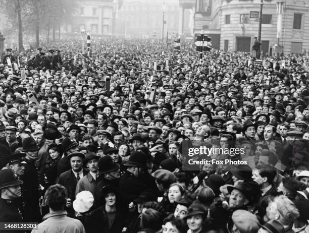 Large crowd of people, some holding cardboard periscopes, gather in Trafalgar Square to catch a glimpse of the British Royals George VI and Queen...