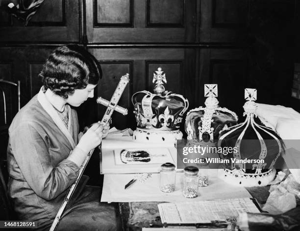 Woman at work on a ceremonial sword, with crowns on the table before her, replicas of the Crown Jewels, merchandise being prepared for the coronation...