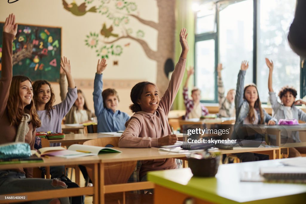 Happy elementary students raising their hands on a class at school.