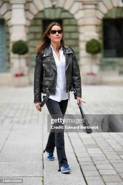 Chloe Street wears black sunglasses, a white shirt, diamonds necklaces, a black faded leather zipper jacket, black denim flared pants, a black shiny...