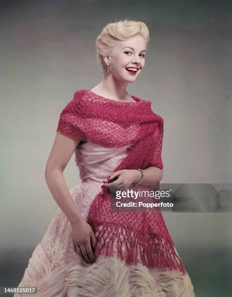Posed studio portrait of English actress Rosalie Ashley wearing a pale pink evening dress with a dark pink crocheted wrap, the dress has a lace...