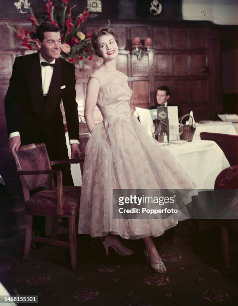 Man holds the chair of his female dining companion as she prepares to be seated at a table to enjoy a dinner date in a restaurant, the woman wears a...
