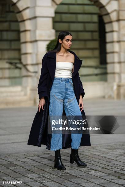 Model wears a black long wool coat, a white latte shoulder-off body, blue denim large pants, black shiny leather block heels ankle boots , outside...
