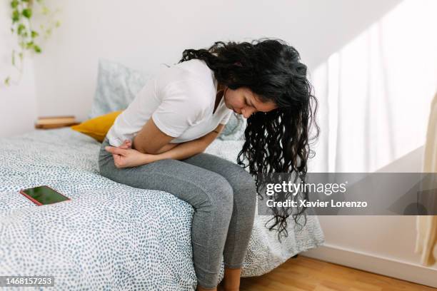 young millennial woman suffering with stomach pain, feeling unwell sitting on bed in the morning - genitalien stockfoto's en -beelden