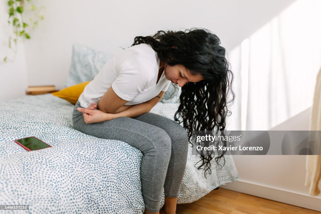 Young millennial woman suffering with stomach pain, feeling unwell sitting on bed in the morning
