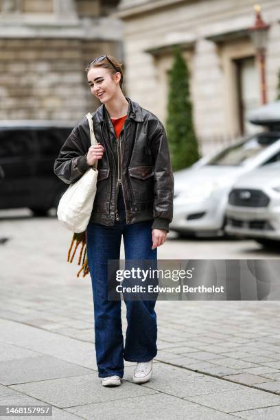 Model wears black glasses, a red t-shirt, a dark brown shiny leather zipper jacket, a white cotton shoulder bag, navy blue denim large pants, white...