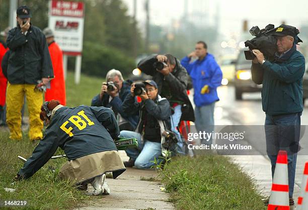 Members of the news media watch as an FBI agent uses a metal detector near the Sunoco gas station October 10, 2002 where a man was shot and killed...