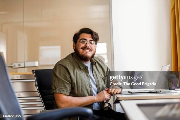 portrait of happy businessman with laptop sitting at desk in office - hot desking arbeitsplatz stock-fotos und bilder