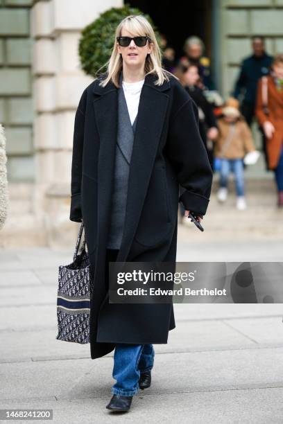 Guest wears black sunglasses, a white t-shirt, a gold necklace, a gray oversized blazer jacket, a black oversized long coat, blue denim large pants,...