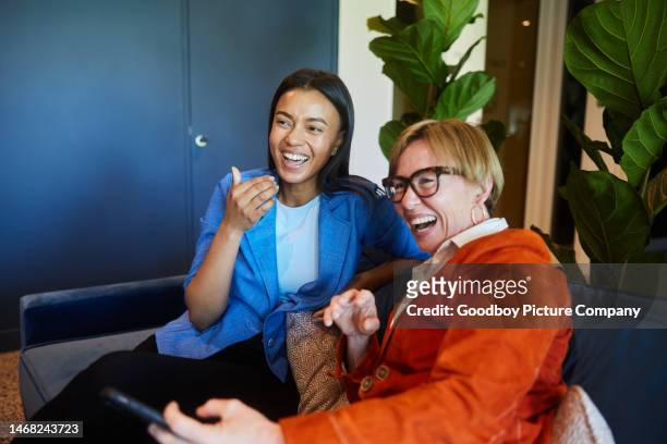 two businesswomen with a phone laughing in an office lounge - coworkers having fun stock pictures, royalty-free photos & images