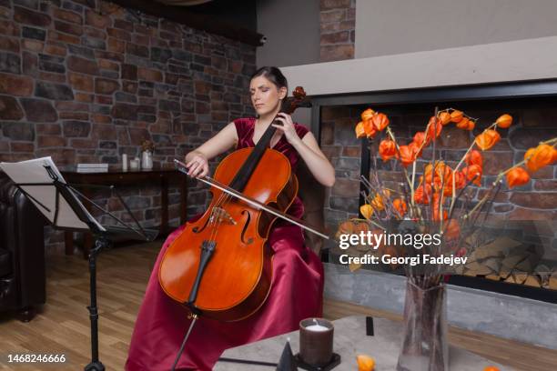 a creative cellist in red dress is playing the cello against a brick wall background and fireplace. - classical musician stock pictures, royalty-free photos & images
