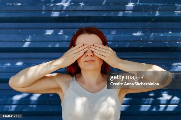 red haired young woman closes her eyes with her palms. concept of three wise monkeys from japanese and indian culture. white girl portrays mizaru, who sees no evil. texture metallic blue background. - 3 wise monkeys stock-fotos und bilder