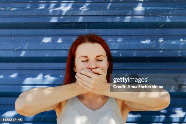 redhead young woman closes her mouth with her hands. concept of three wise monkeys from japanese and indian culture. white female portrays iwazaru, who speaks no evil. textured blue background. - 3 wise monkeys stock-fotos und bilder