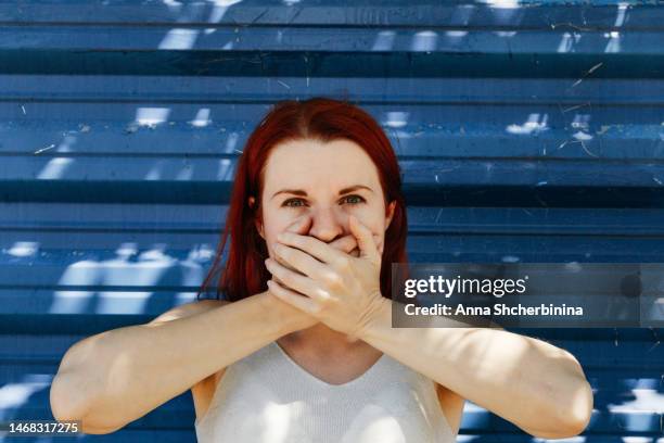 redhead young woman closes her mouth with her hands. concept of three wise monkeys from japanese and indian culture. white girl portrays iwazaru, who speaks no evil. textured metallic blue background. - 3 wise monkeys stock-fotos und bilder