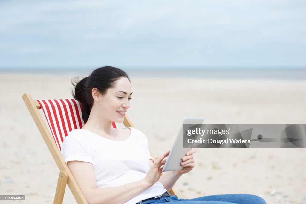 Woman sitting on deck chair at beach with tablet.