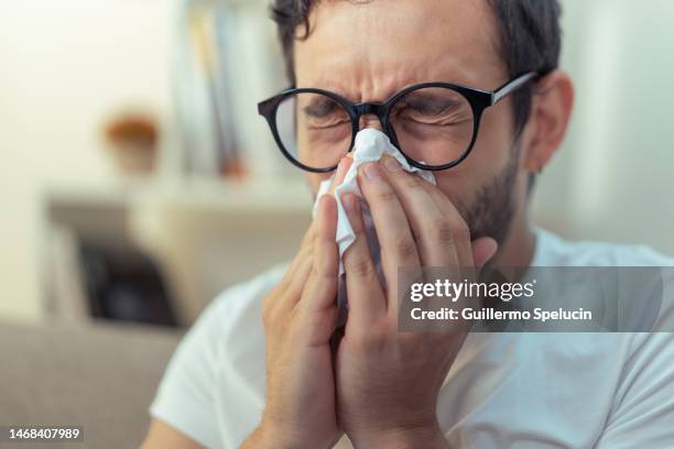 young man sneezing, wiping his nose with a piece of tissue paper - allergie foto e immagini stock