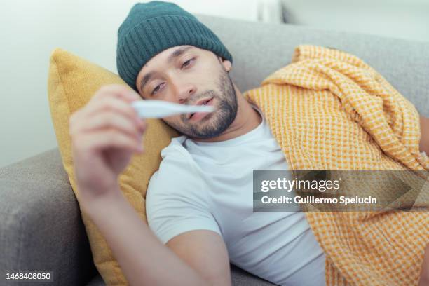 young man with fever taking his temperature, lying on the living room couch. - fever fotografías e imágenes de stock