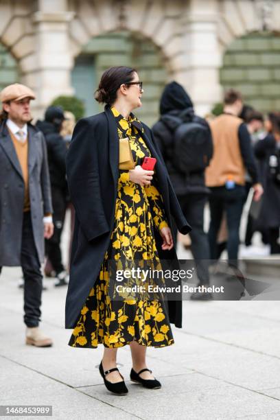 Guest wears black sunglasses, a black and yellow flower print pattern midi dress, a black long jacket, a beige shiny leather shoulder bag, black...
